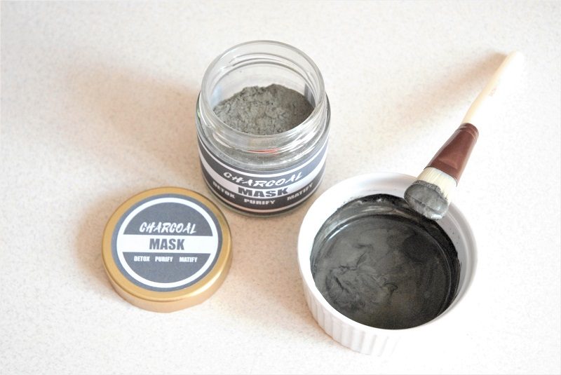 dry mix of DIY charcoal mask in a jar with a dark grey charcoal printable label on the jar. Next to it a bowl with a dry charcoal mask mixed with water and a brush